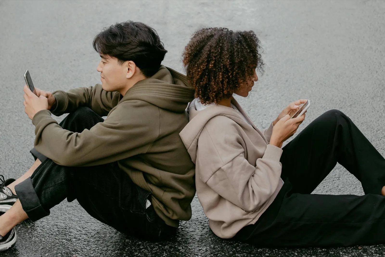 Two young people sitting back to back on asphalt, each absorbed in their own phone