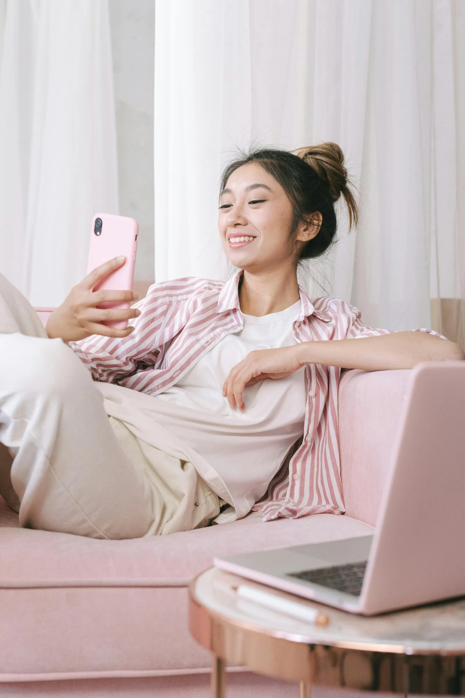 Young woman smiling at her phone while relaxing on a pink sofa, an open laptop on the table beside her