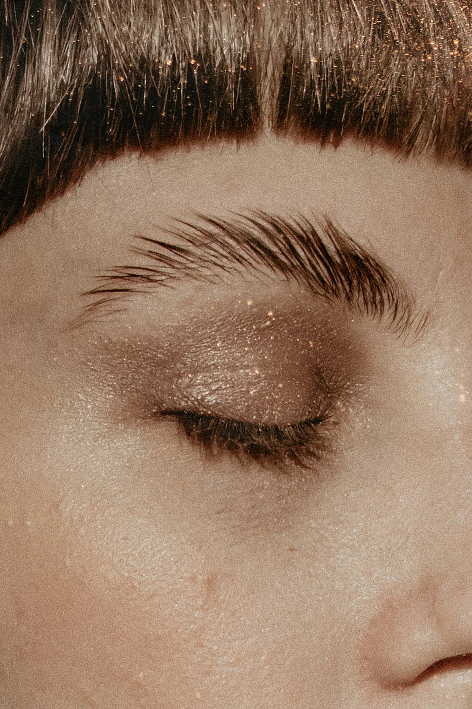 Extreme close-up of a closed eye with gold glitter eyeshadow across the lid, the same glitter scattered through a blunt fringe above